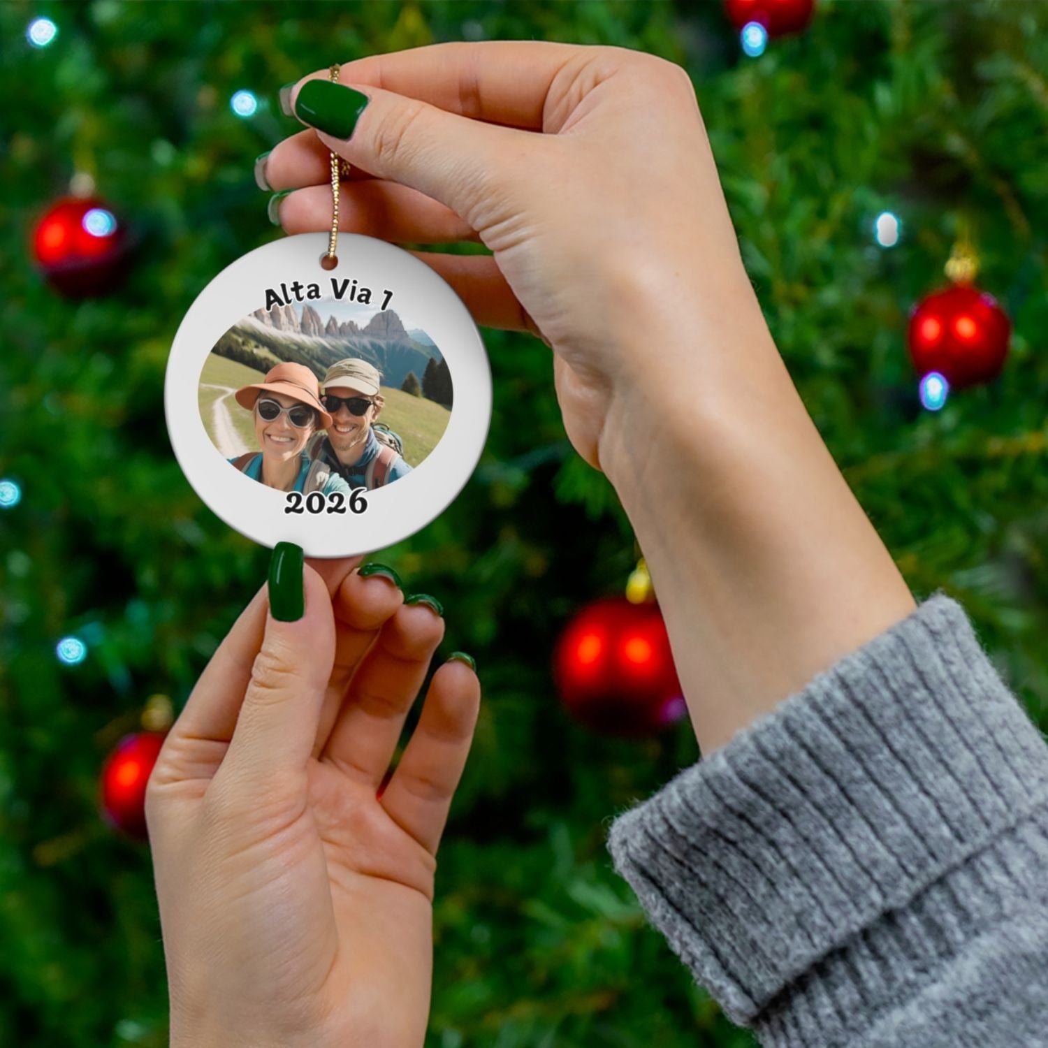 Person holding a personalized Alta Via 1 ornament with custom photo and year in front of a decorated Christmas tree, highlighting the option to upload your own hiking picture and showcasing the size of the ornament.