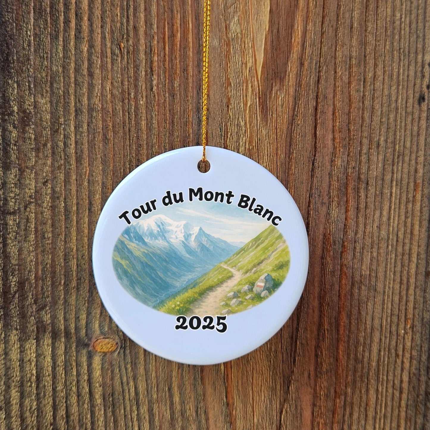 Close-up of Tour du Mont Blanc 2025 ceramic ornament with watercolor mountain trail design, hanging on rustic wooden background.