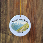 Close-up of Tour du Mont Blanc 2025 ceramic ornament with watercolor mountain trail design, hanging on rustic wooden background.